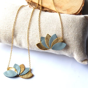 Peut inclure: Deux colliers en chaîne dorée avec un pendentif en forme de fleur de lotus en cuir gris et or. Les colliers sont présentés sur une surface en tissu blanc.