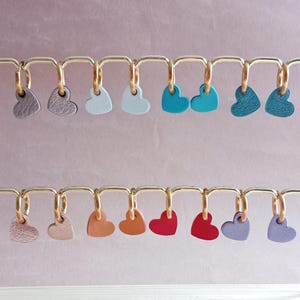Pode incluir: Uma exibição de 12 pingentes de coração de couro em várias cores, incluindo roxo, cinza, azul, vermelho e laranja. Os pingentes estão pendurados em argolas douradas em uma barra de metal dourada.