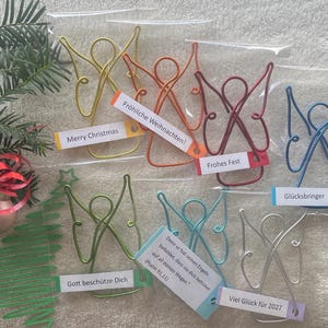 Könnte beinhalten: Sieben Drahtengel in Gold, Orange, Rot, Blau, Grün, Hellblau und Silber, jeweils in einer transparenten Kunststoffhülle. Jeder Engel hat ein Papieretikett mit Text, darunter Merry Christmas, Fröhliche Weihnachten und Viel Glück für 2027.