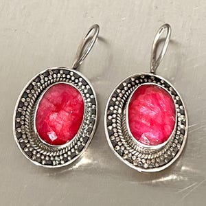 May include: A pair of oval-shaped earrings featuring vibrant red gemstones set in a silver-toned metal. The earrings have a detailed border with small, dark accents, and a simple hook closure.