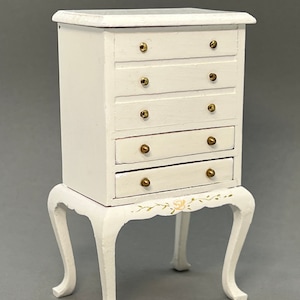 May include: White wooden chest of drawers with five drawers and gold knobs. The chest is on four cabriole legs and has a floral design painted on the bottom.