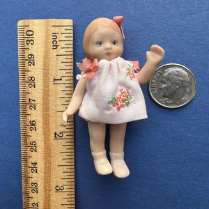 May include: A miniature porcelain doll with light brown hair, wearing a pale pink dress with small floral embroidery and matching bows. The doll stands approximately 2.5 inches tall next to a wooden ruler and a United States dime for scale.