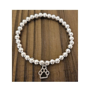 May include: Silver beaded bracelet with a paw print charm. The bracelet is made of small, round, shiny silver beads. A silver charm shaped like a dog's paw hangs from the centre. The bracelet is shown on a wooden surface.