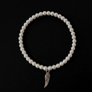 May include: A white pearl bracelet with a silver wing charm. The bracelet is made of round, white pearls and hangs against a black background. The wing charm is detailed and adds a delicate touch to the jewelry.