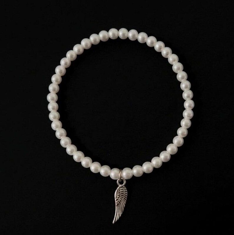 May include: A white pearl bracelet with a silver wing charm. The bracelet is made of round, white pearls and hangs against a black background. The wing charm is detailed and adds a delicate touch to the jewelry.