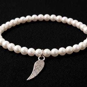May include: White beaded bracelet with a silver wing charm. The bracelet features a strand of round, white beads. The charm is a detailed silver wing. The bracelet is displayed against a black background.