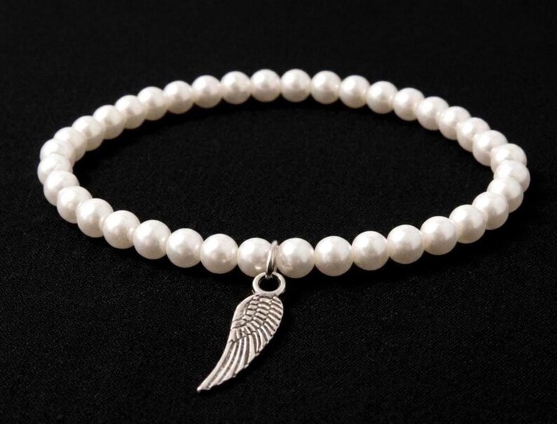 May include: White beaded bracelet with a silver wing charm. The bracelet features a strand of round, white beads. The charm is a detailed silver wing. The bracelet is displayed against a black background.