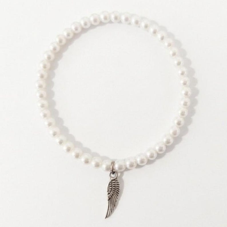 May include: A white pearl bracelet with a silver-colored wing charm. The bracelet is made of small, round, white pearls strung together. The wing charm is detailed and hangs from the center of the bracelet.