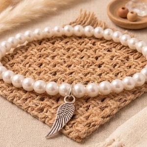 May include: A white pearl bracelet with a silver wing charm. The bracelet is displayed on a woven, brown square. The background includes a small wooden bowl with decorative items and dried plants.