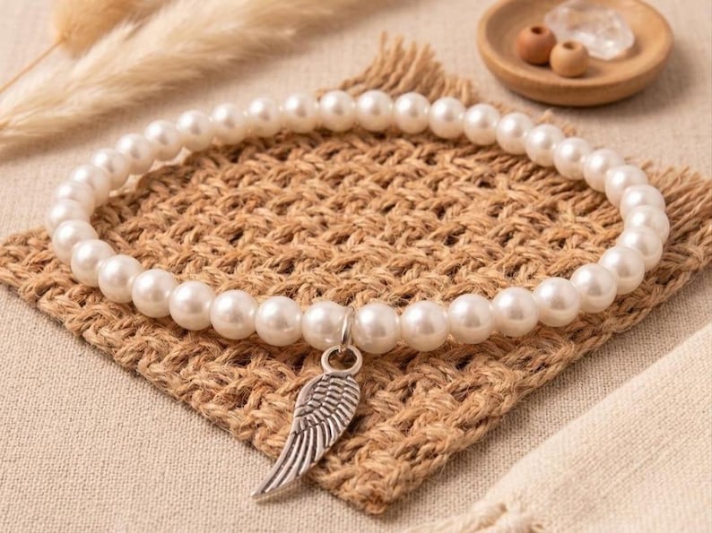 May include: A white pearl bracelet with a silver wing charm. The bracelet is displayed on a woven, brown square. The background includes a small wooden bowl with decorative items and dried plants.