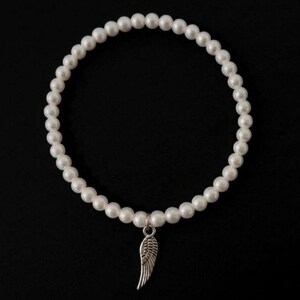 May include: White pearl bracelet with a silver-colored wing charm. The bracelet is made of round, white pearls strung together. The wing charm is detailed and hangs from the center of the bracelet against a black background.