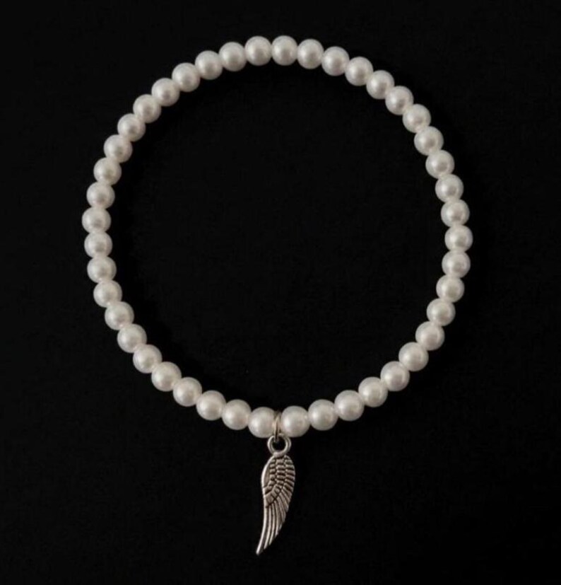 May include: White pearl bracelet with a silver-colored wing charm. The bracelet is made of round, white pearls strung together. The wing charm is detailed and hangs from the center of the bracelet against a black background.