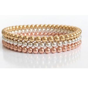 May include: Three beaded bracelets stacked together. The top bracelet is gold, the middle is silver, and the bottom is rose gold. The bracelets are made of small, round beads and are arranged on a white surface.