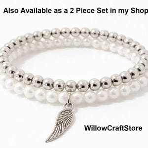 May include: Two beaded bracelets. One bracelet features silver-colored beads, and the other has white pearl-like beads. A silver-colored wing charm hangs from the bracelets. The bracelets are stacked on top of each other.