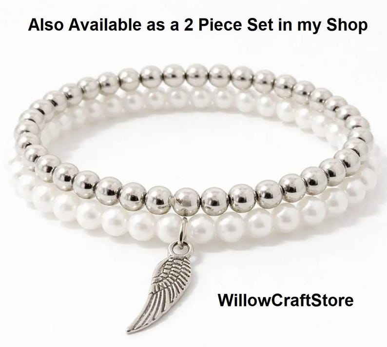 May include: Two beaded bracelets. One bracelet features silver-colored beads, and the other has white pearl-like beads. A silver-colored wing charm hangs from the bracelets. The bracelets are stacked on top of each other.