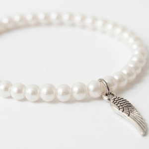 May include: White pearl bracelet with a silver-colored wing charm. The bracelet is made of round, white pearls strung together. The wing charm is detailed and hangs from a small ring attached to the bracelet.