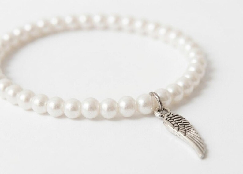 May include: White pearl bracelet with a silver-colored wing charm. The bracelet is made of round, white pearls strung together. The wing charm is detailed and hangs from a small ring attached to the bracelet.