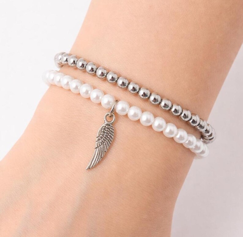 May include: Two beaded bracelets. One bracelet features small, silver-colored beads. The other bracelet has white, pearl-like beads and a silver-colored wing charm. The bracelets are worn on a wrist.