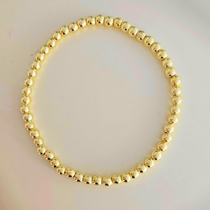 May include: A gold beaded bracelet. The bracelet is made of small, round, gold-coloured beads strung together. The beads have a shiny, reflective surface. The bracelet is displayed on a white surface.