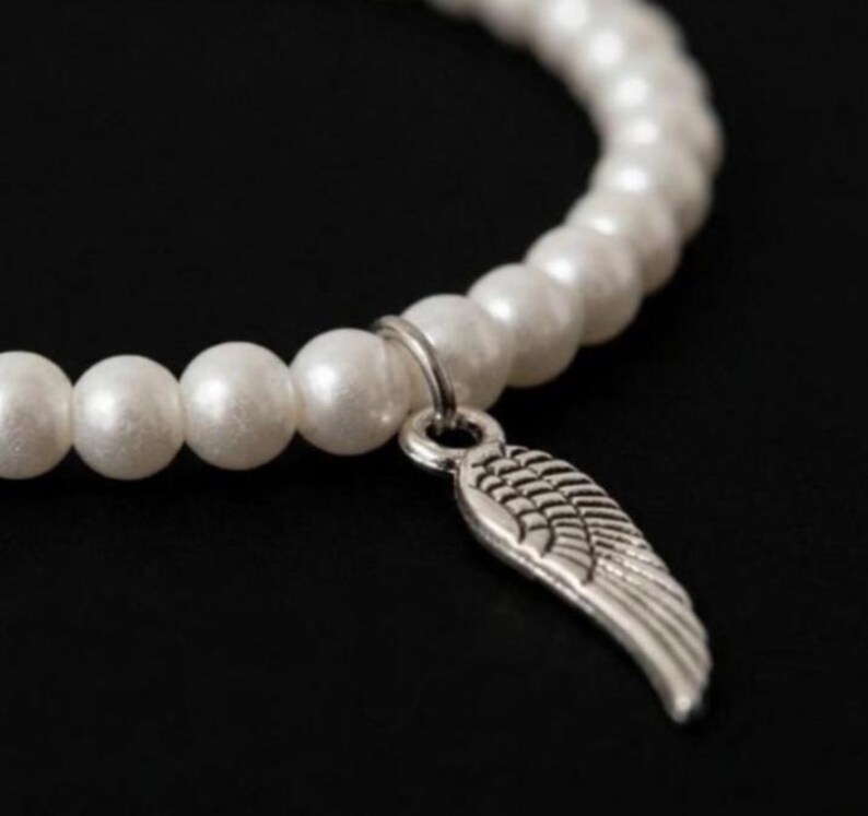 May include: A white pearl bracelet with a silver wing charm. The bracelet features a strand of round, lustrous white pearls. The silver charm is a detailed wing design, adding a touch of elegance to the jewelry.