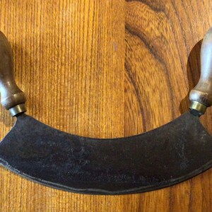 May include: A curved mezzaluna knife with wooden handles. The blade is dark metal and has a slight curve. The handles are light brown and attached to the blade with brass-coloured metal rings. The knife rests on a wooden surface.
