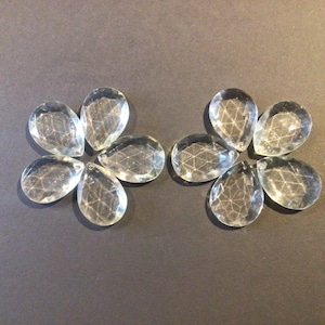May include: Two sets of clear, faceted teardrop-shaped crystal beads arranged in a flower-like pattern. The beads have internal geometric patterns and are displayed against a dark grey background.