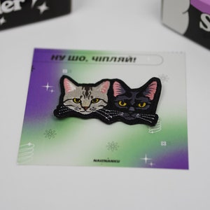 May include: A colourful patch featuring two cat faces, one grey tabby and one black cat, with the text "Ну шо, чипляй!" in Cyrillic script.