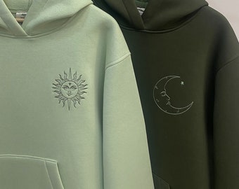 May include: Two hooded sweatshirts hanging on a rack. The sweatshirt on the left is light green with a sun graphic. The sweatshirt on the right is dark green with a crescent moon graphic.