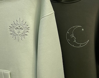May include: Two hooded sweatshirts, one light green with a sun and the other dark green with a crescent moon, both with embroidered designs.