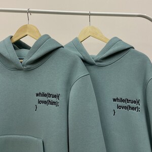 May include: Two light blue hooded sweatshirts with black text. The text on the left sweatshirt reads "while(true){ love(him);}". The text on the right sweatshirt reads "while(true){ love(her);}".
