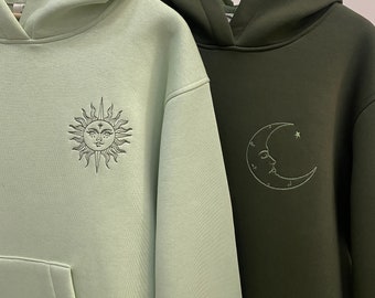 May include: Two hooded sweatshirts, one light green with a sun and the other dark green with a crescent moon, both with embroidered designs.