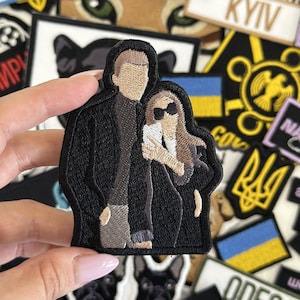 May include: Embroidered patch featuring a couple, a man and a woman, standing side-by-side. The man is wearing a black jacket and the woman is wearing a black dress. The patch is surrounded by other patches, including a Ukrainian flag.