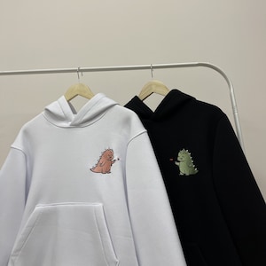 Peut inclure: Deux sweat-shirts &agrave; capuche avec des motifs de dinosaures. Le sweat-shirt de gauche est blanc avec un dinosaure rose tenant un c&oelig;ur rouge. Le sweat-shirt de droite est noir avec un dinosaure vert tenant un c&oelig;ur rouge.