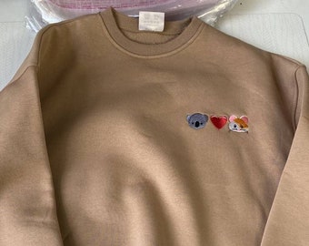 Puede incluir: Una sudadera marrón con un pequeño diseño bordado de un koala, un corazón y un ratón. La sudadera tiene cuello redondo y mangas largas.