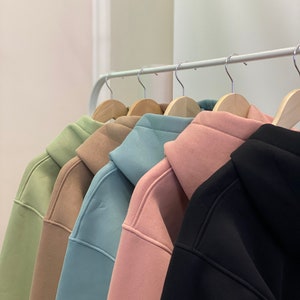 May include: Five hooded sweatshirts hanging on a rack. The sweatshirts are in different colours: green, brown, light blue, pink, and black.