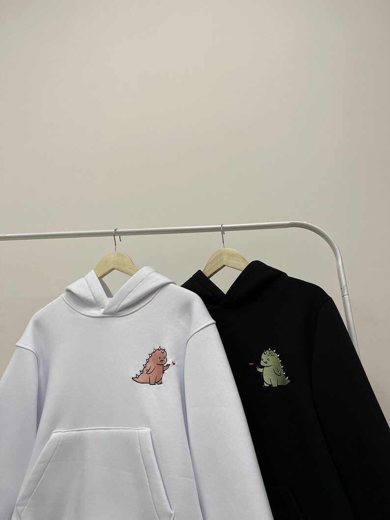 Peut inclure: Deux sweat-shirts &agrave; capuche avec des motifs de dinosaures. Le sweat-shirt de gauche est blanc avec un dinosaure rose tenant un c&oelig;ur rouge. Le sweat-shirt de droite est noir avec un dinosaure vert tenant un c&oelig;ur rouge.