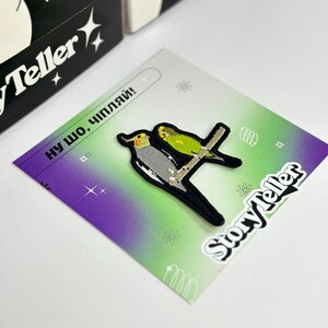 May include: A green and purple background with a white sticker that says "Storyteller" on it. The sticker is over a black and white embroidered patch of two birds on a branch. The text "ну шо, чипляй!" is printed on the background.