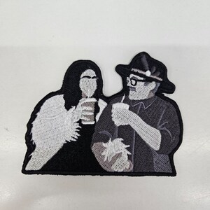 Peut inclure: Patch brodé représentant deux personnages, une femme aux longs cheveux noirs et un homme portant un chapeau et des lunettes, tous deux tenant des tasses. Le patch est noir avec des broderies blanches et grises.
