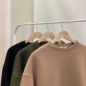 Peut inclure: Trois sweats &agrave; capuche suspendus &agrave; des cintres en bois. Les sweats sont noir, vert et beige. Le sweat beige porte l'inscription "ARTISAN&CO" imprim&eacute;e sur le cintre.