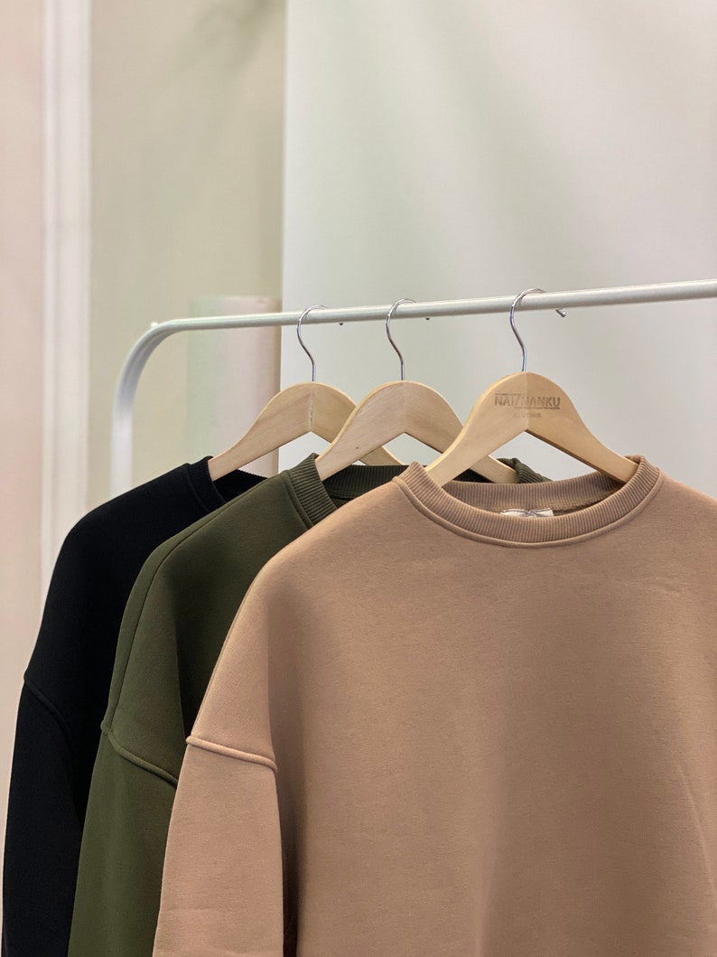 Peut inclure: Trois sweats &agrave; capuche suspendus &agrave; des cintres en bois. Les sweats sont noir, vert et beige. Le sweat beige porte l'inscription "ARTISAN&CO" imprim&eacute;e sur le cintre.