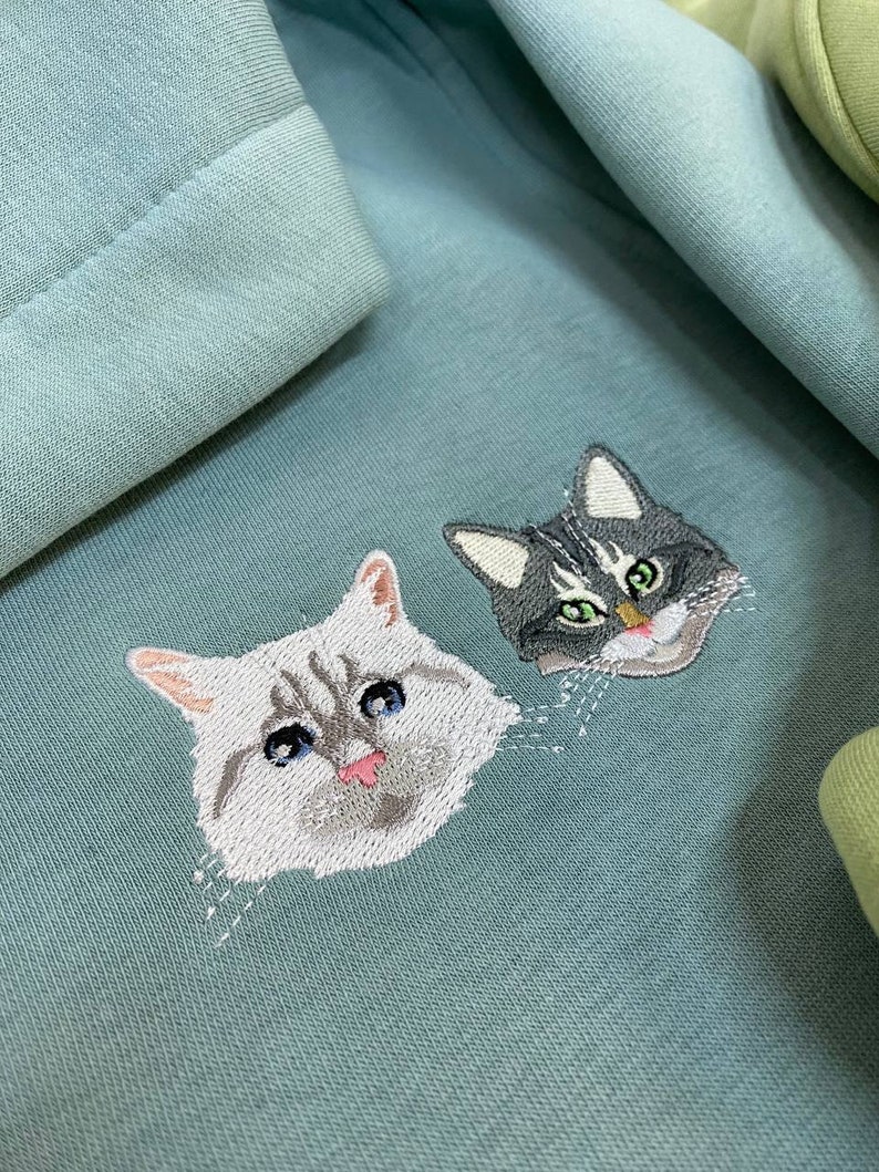 May include: A teal blue sweatshirt with two embroidered cat faces. The cat on the left is white with blue eyes and a pink tongue. The cat on the right is grey with green eyes.