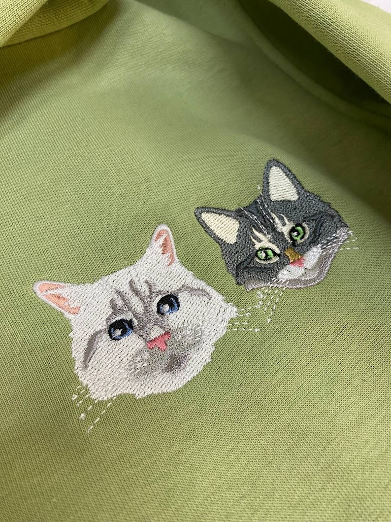 May include: A green fabric with two embroidered cat faces. The cat on the left has white fur with grey markings and blue eyes. The cat on the right has grey fur with white markings and green eyes.