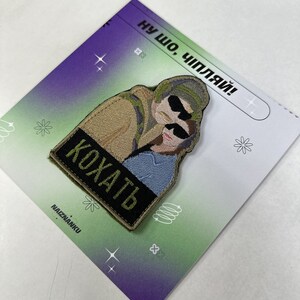 May include: A green and black embroidered patch with the text "кохать" in Cyrillic script. The patch features a cartoon image of two people wearing sunglasses and jackets. The text "ну шо, чипляй!" is printed on a white background.