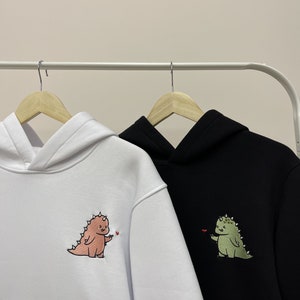 Peut inclure: Deux sweat-shirts &agrave; capuche avec des motifs de dinosaures brod&eacute;s. Un sweat-shirt est blanc avec un dinosaure rose, l'autre est noir avec un dinosaure vert.