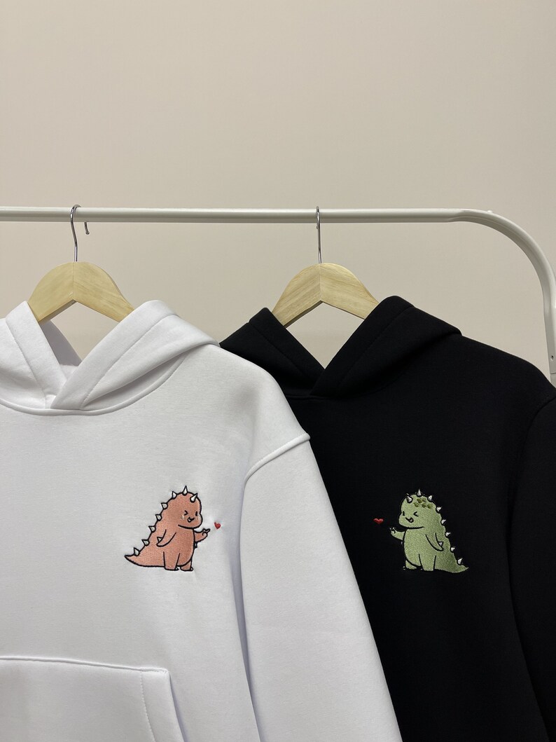 Peut inclure: Deux sweat-shirts &agrave; capuche avec des motifs de dinosaures brod&eacute;s. Un sweat-shirt est blanc avec un dinosaure rose, l'autre est noir avec un dinosaure vert.