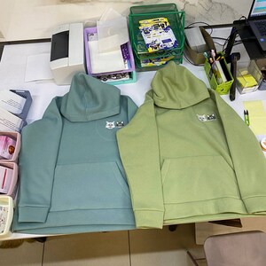 May include: Two hooded sweatshirts, one teal green and one light green, with a small white cat graphic on the front. Both sweatshirts have a kangaroo pocket.