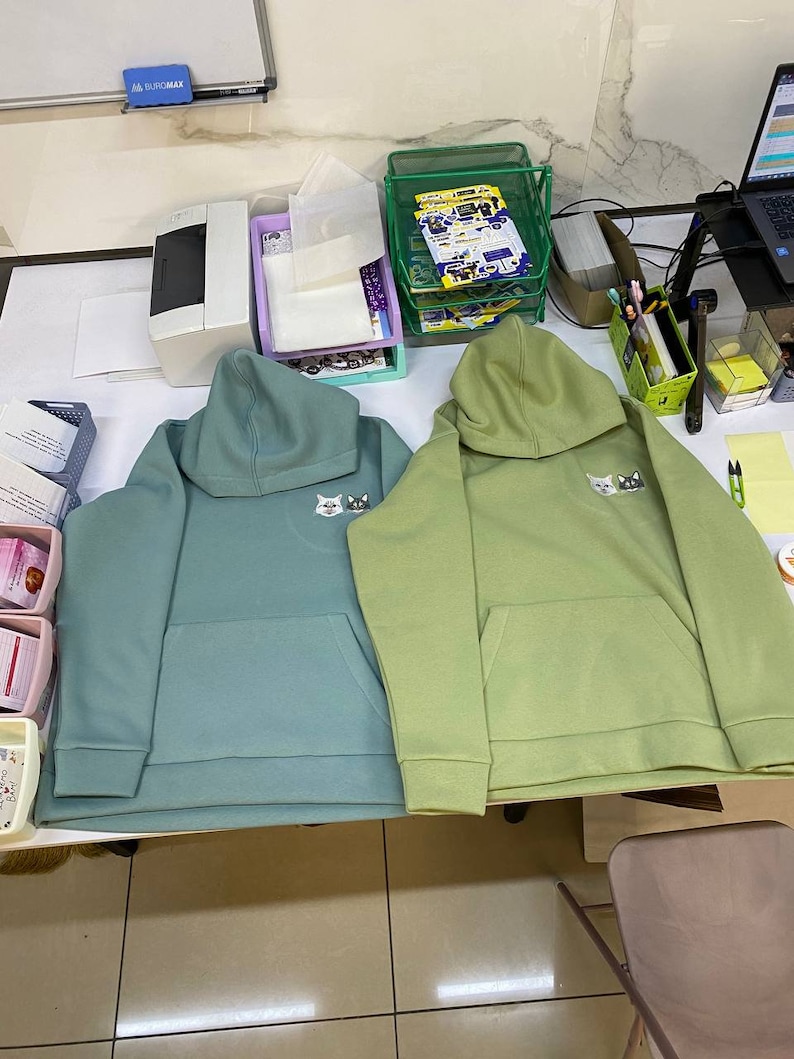 May include: Two hooded sweatshirts, one teal green and one light green, with a small white cat graphic on the front. Both sweatshirts have a kangaroo pocket.