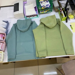 May include: Two hooded sweatshirts, one teal green and one light green, laid out on a white table.
