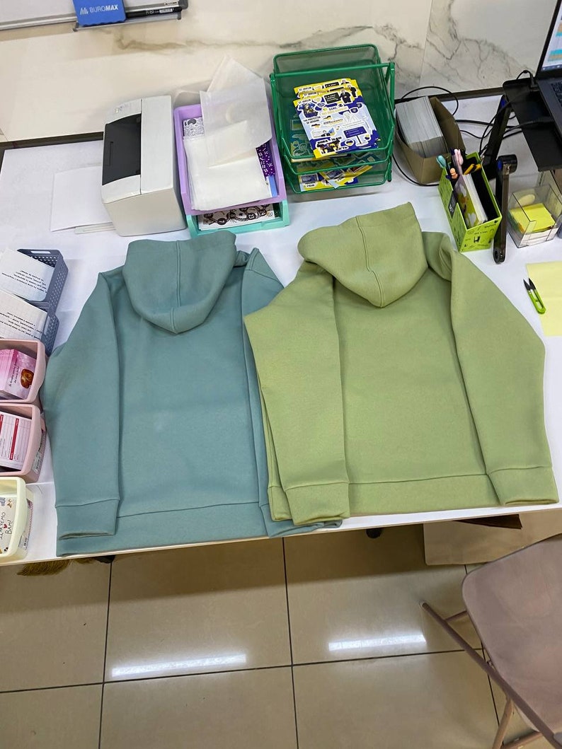 May include: Two hooded sweatshirts, one teal green and one light green, laid out on a white table.