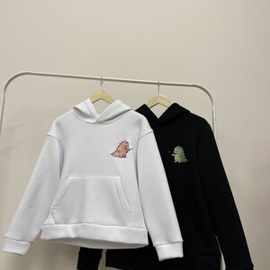 Peut inclure: Deux sweat-shirts &agrave; capuche avec des motifs de dinosaures. L'un est blanc avec un dinosaure rose et l'autre est noir avec un dinosaure vert.